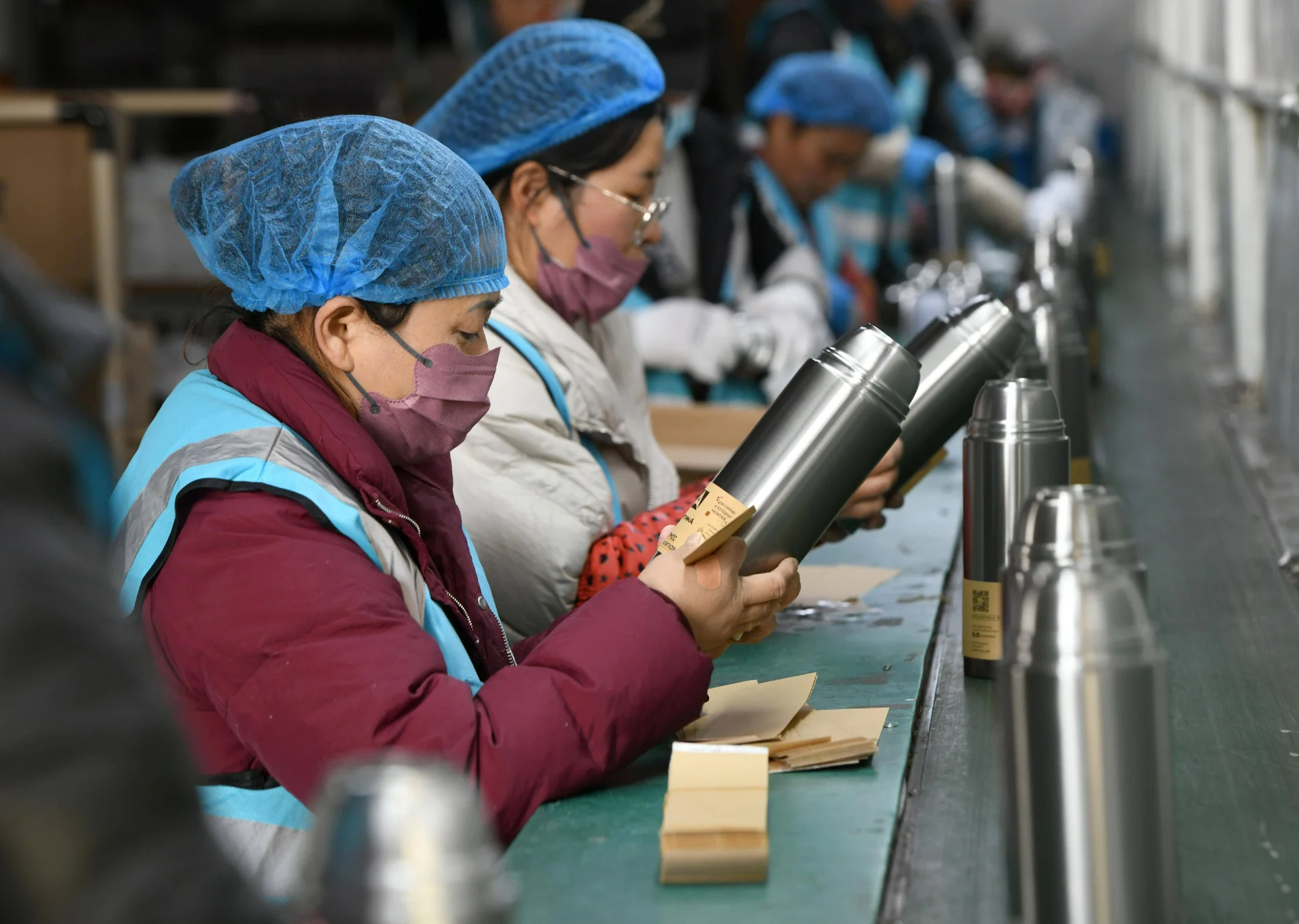 Factory workers wearing blue hair nets inspecting stainless steel thermoses on a production line.