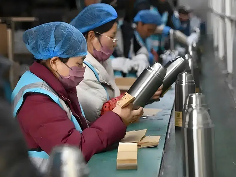 Workers inspecting stainless steel cups on an assembly line wearing blue caps and safety attire.